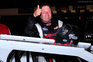 Ein Mann in einem schwarzen und weißen Rennanzug und Helm steht neben einem weißen Fahrzeug und gibt einen Daumen hoch, mit einem anderen Mann in einem roten und schwarzen Rennanzug links von ihm und einem schwarzen Fahrzeug und Tafeln im Hintergrund.