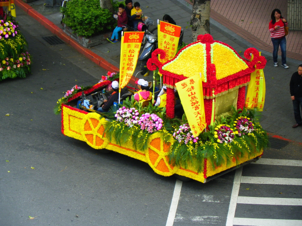 Schwimmend in einem mit Blumen und Bannern geschmückten Paradewagen mit Reitern, mit Zuschauern auf der rechten Seite und Bäumen im Hintergrund.
