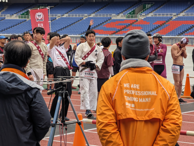 Eine Gruppe von Menschen auf einer Bahn mit einer Kamera auf einem Stativ im Vordergrund, Stühle, ein Banner und Scheinwerfer im Hintergrund.