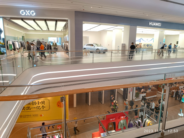 Das Innere eines Einkaufszentrums mit Menschen, die herumlaufen, Glasgeländern, Kleidung an Bügeln, Texttafeln, Säulen, Deckenleuchten und einem geparkten Auto im Hintergrund, mit einem GXG und Huawei-Partnerschafts-Wasserzeichen unten.
