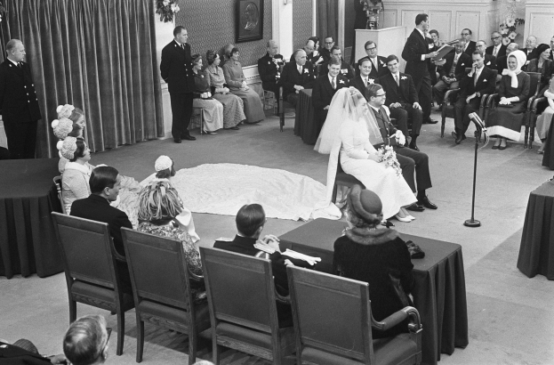 Schwarz-weiß-Foto einer Hochzeitszeremonie mit sitzenden und stehenden Menschen, einem Mann mit Mikrofon und einer Frau mit Blumenstrauß, mit einem Podium mit Mikrofon, Blumenschmuck, einem Vorhang, einem gerahmten Foto an der Wand und einer Tür im Hintergrund.