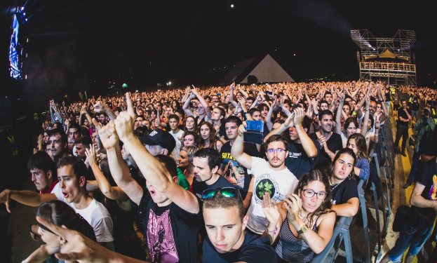 Eine große Menge Menschen auf einem Musikfestival mit erhobenen Händen, einige sitzen auf Stühlen und andere stehen, begrenzt von einem Metallzaun auf der rechten Seite, mit Gebäuden und Lichtern im Hintergrund unter einem dunklen Himmel.