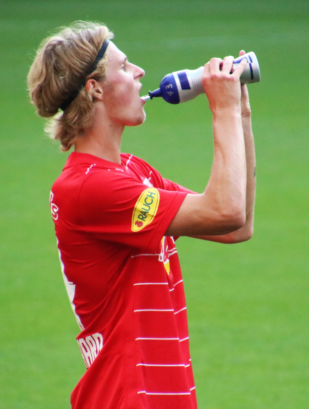 Ein Mann in einem roten T-Shirt trinkt aus einer Flasche, während er auf einem Fußballfeld steht.