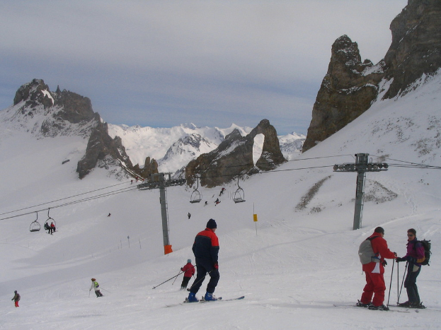 Menschen in Pullovern fahren auf der Eisbahn Ski, mit einer Seilbahn, Bergen und einem bewölkten Himmel im Hintergrund.