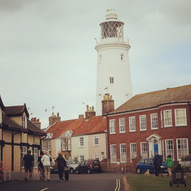 Eine Straßenszene mit Gebäuden, parkenden Autos, Menschen, die gehen, Hunden, einem Objekt auf dem Gras und einem Turm unter dem Himmel.