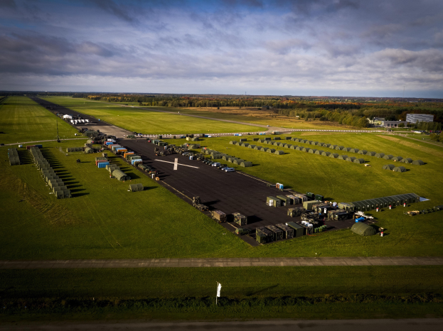 Luftaufnahme einer Militärbasis in den Niederlanden mit Fahrzeugen, Gras, Bäumen, Gebäuden und einer bewölkten Himmel.
