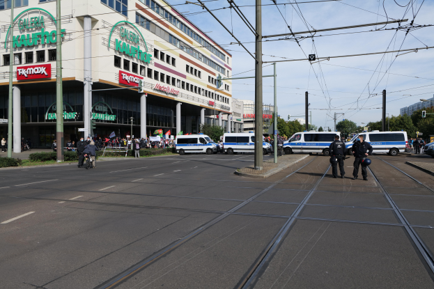 Eine Gruppe von Polizeißen, die auf der Straßenseite neben einer Tram stehen, mit Fahrzeugen, Strommasten, Gebäuden mit Namensschildern, Bäumen und einem bewölktem Himmel im Hintergrund.