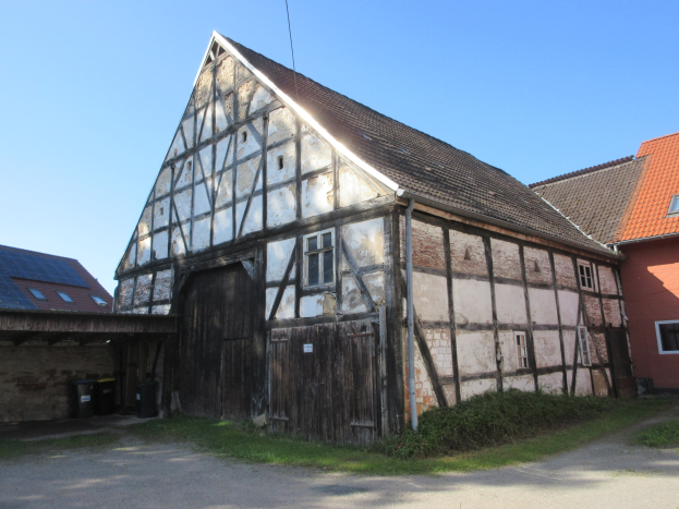 Altes Gebäude mit einer hölzernen Tür und Fenstern, umgeben von Gras und Pflanzen, Solarpaneele auf dem Dach, klarer blauer Himmel im Hintergrund.