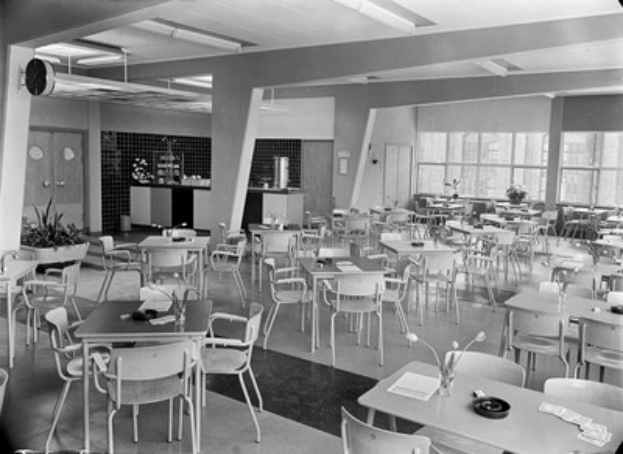 Schwarz-weißes Foto einer Cafeteria mit Tischen und Stühlen, Tellern, Besteck, Gläsern und Blumenvasen auf den Tischen sowie Türen, Fenstern und Deckenleuchten im Hintergrund.