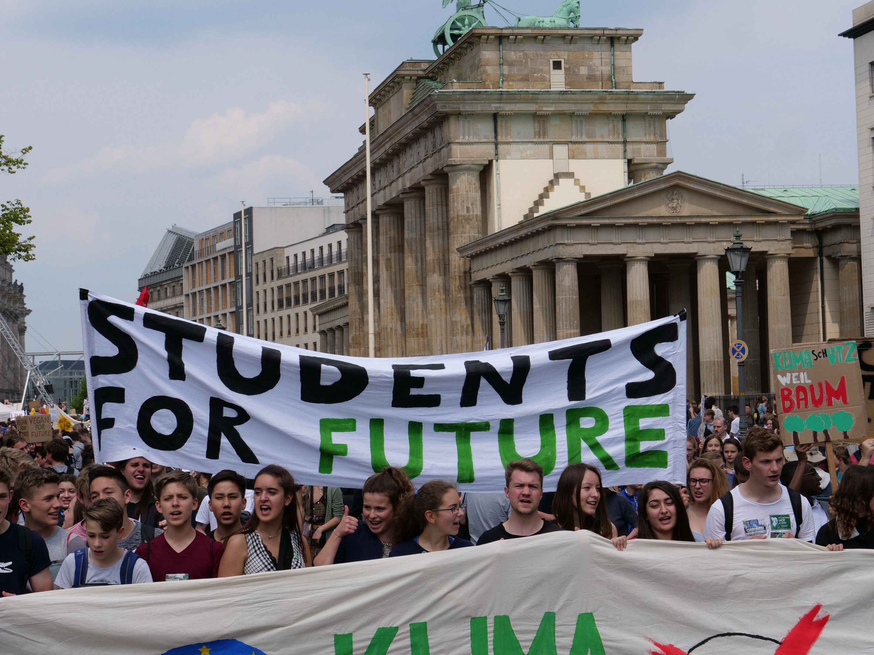 Eine Gruppe von Schülern marschiert in Berlin, eine bunt bemalte "Schüler für die Zukunft"-Fahne schwingend, vor einer Kulisse aus Gebäuden, Bäumen und Himmel.