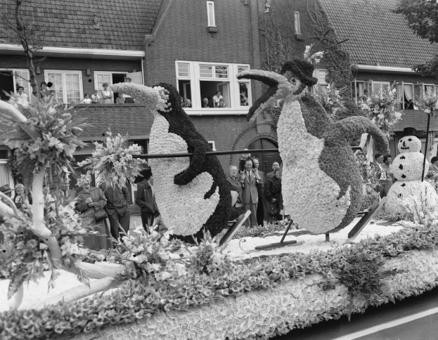 Schwarzes und weißes Foto eines Umzugswagens mit Blumen und Pinguinfiguren, mit Zuschauern, Bäumen und Gebäuden im Hintergrund.