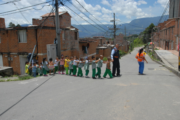 Zwei Kinder, ein erwachsener Mann und eine erwachsene Frau überqueren eine Straße in einer Reihe, mit Gebäuden auf beiden Seiten, Pfählen, Drähten, Bäumen, Hügeln und einem bewölkten Himmel im Hintergrund.