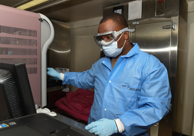 Ein Mann in einem blauen Laborkittel und Schutzbrille arbeitet an einem Computer, umgeben von verschiedenen Gegenständen, mit einer Wand voller Papiere im Hintergrund, was darauf hindeutet, dass er Teil eines Ebola-Forschungsteams ist.
