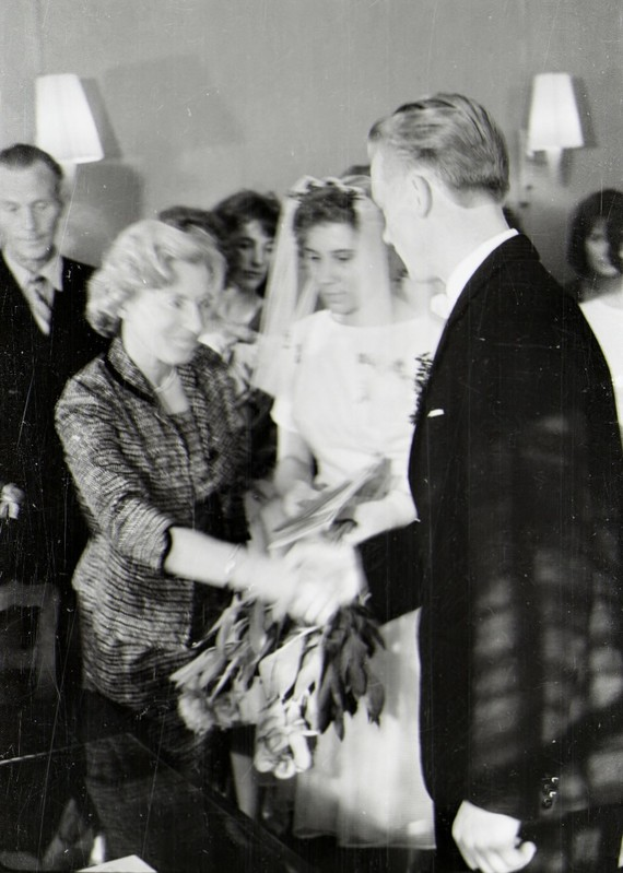 Ein Schwarz-Weiß-Foto eines Brautpaares beim Austausch der Trauringe, wobei die Braut in einem weißen Kleid und Schleier mit einem Strauß Blumen in der Hand zu sehen ist und der Bräutigam in einem Anzug und Krawatte steht, mit Wandlampen im Hintergrund.