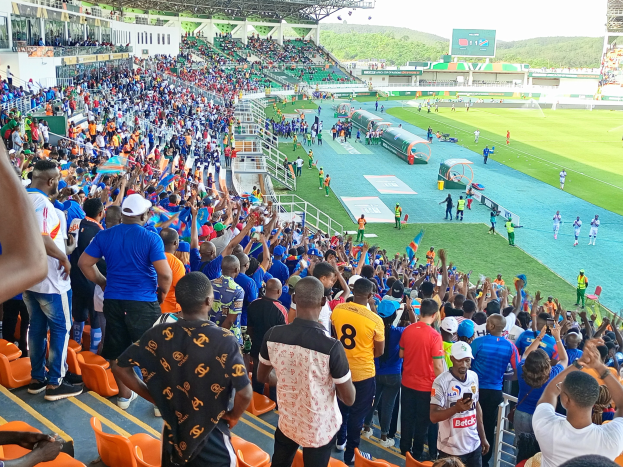 Eine große Menschenmenge versammelt sich vor einem eingezäunten Fußballfeld mit Hügeln und einem klaren blauen Himmel im Hintergrund und erwartet den Beginn eines Spiels während der Afrikameisterschaft 2019.