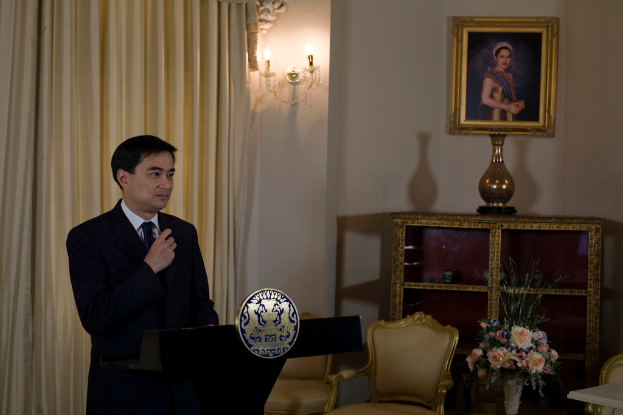 Eine Person steht an einem Rednerpult mit einem Vorhang dahinter, einer Blumenvase und Stühlen rechts und gerahmten Bildern mit daran befestigten Lampen an der Wand.