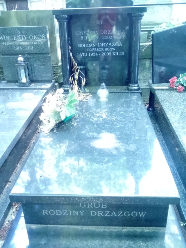 Ein Friedhof mit zwei benachbarten Grabsteinen, umgeben von Blumenbouquets und anderen Gegenständen, mit einem Gebäude im Hintergrund.