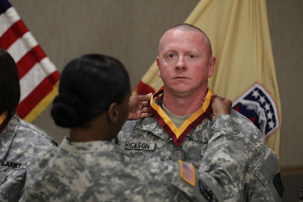 Eine Frau in Armeeuniform berührt die Medaille eines Mannes, der vor ihr steht, mit sichtbaren Flaggen im Hintergrund.