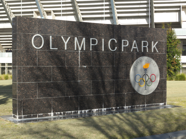 Der Olympische Park in Melbourne, Australien, mit einem Namensschild im Vordergrund, Gras, Bäumen, Pflanzen und einem Gebäude in der Ferne.