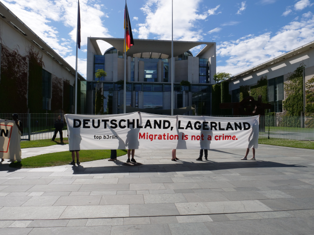 Eine Gruppe von Menschen hält ein Transparent mit der Aufschrift "Deutschland, Lagerland Migration ist kein Verbrechen" vor einem Gebäude mit Fenstern, umgeben von Gras, einem Metallzaun, Pflanzen, Bäumen und Fahnenmasten, unter einem bewölkten Himmel.