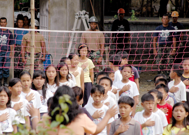 Kinder vor einem Netz mit einer Frau im Vordergrund, Menschen mit Helmen, einer Leiter, einer Wand und Bäumen im Hintergrund.