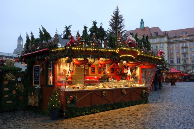 Ein Weihnachtsmarktstand auf einem Kopfsteinpflasterweg, umgeben von Gebäuden, geschmückten Bäumen und Menschen, mit einem sichtbaren Himmel im Hintergrund.