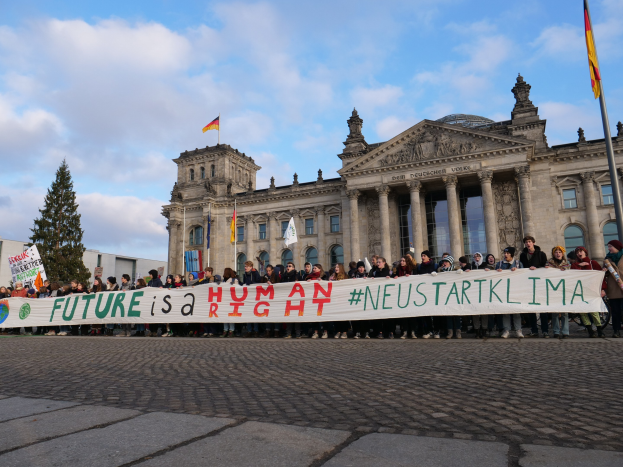 Gruppe von Menschen vor dem Reichstagsgebäude in Berlin mit einer Fahne, auf der 'Zukunft ist ein Menschenrecht' steht, mit Säulen, Fenstern und Bögen des Gebäudes im Hintergrund, umgeben von Bäumen und Fahnenmasten bei bewölktem Himmel.