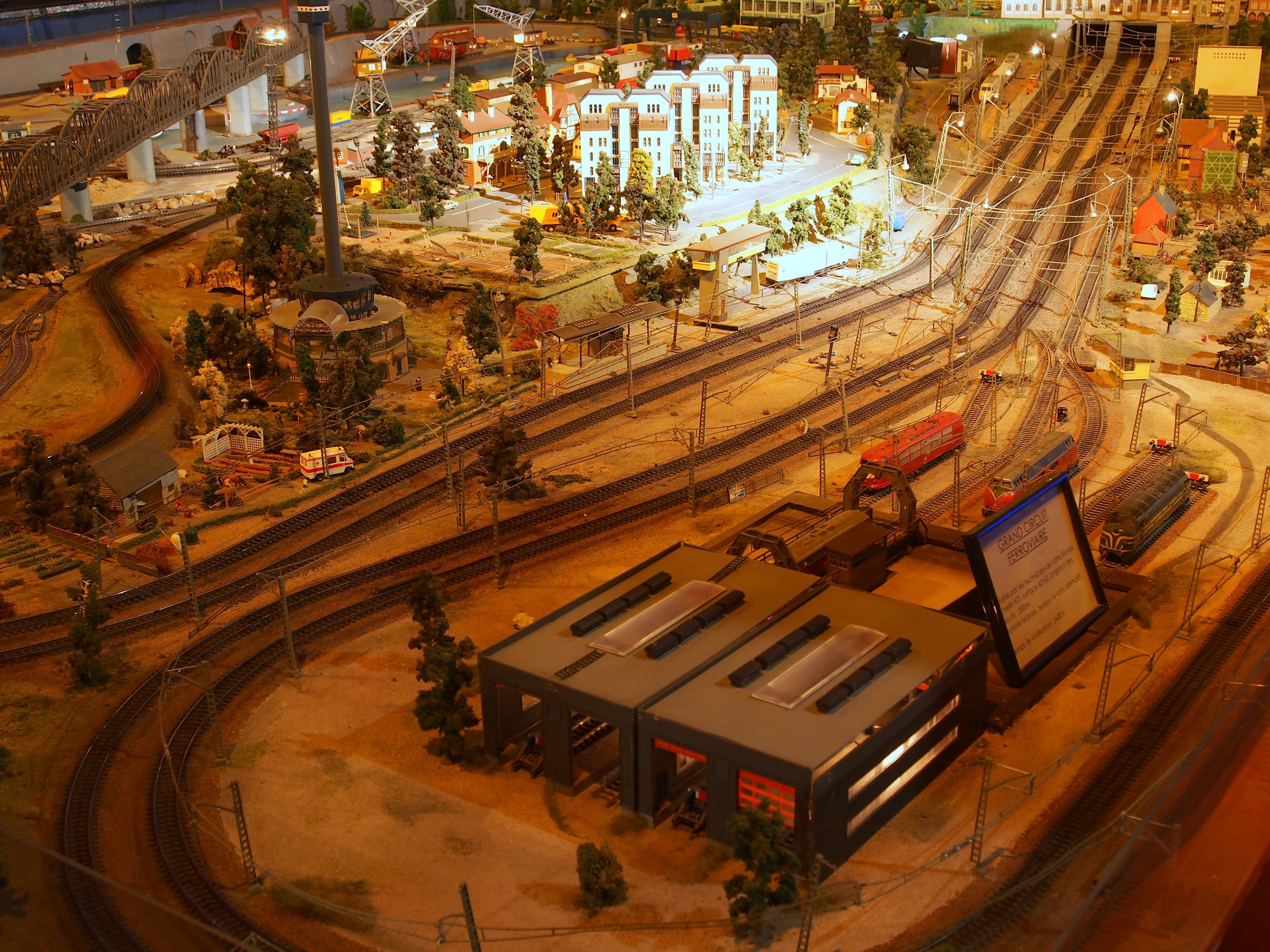 Eine detaillierte Modellbahnausstellung in einem Museum, mit Gebäuden, Bäumen, Schienen, Fahrzeugen, Pfählen, Lichtern und Plakaten, die eine belebte Stadtlandschaft nachahmen.