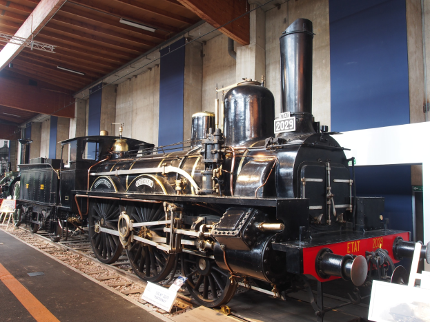 Alte Dampflokomotive in einem Museums Setting mit umgebenen Eisenbahnschienen, Informationsschildern und Beleuchtungselementen.