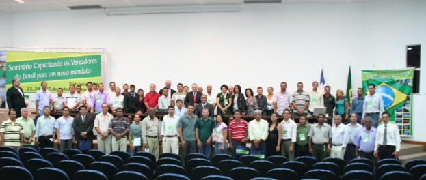 Ein geräumiger Saal mit ein paar Menschen, die für ein Bild posieren, leere Stühle im Vordergrund, zwei Flaggen und zwei Banner im Hintergrund und zwei Lautsprecher auf der rechten Seite, mit Beleuchtung oben.