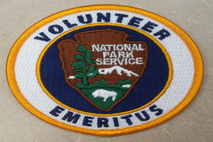 Ein rundes Patch mit einem blauen Rand, weißem Hintergrund und fettem schwarzem Text, der 'Volunteer National Park Service Emeritus' lautet und in der Mitte eines gelben Kreises angeordnet ist.