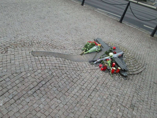 Gedenkstätte mit Blumen, Kerzen und verstreuten Buketts am Straßenrand, umgeben von Bäumen, Gebäuden und Fahrzeugen mit angeketteten Pfählen.