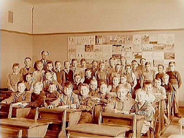 Schwarz-weißes Foto einer Klasse mit Kindern auf Bänken sitzend und stehend, Papiere an der Rückwand angebracht.