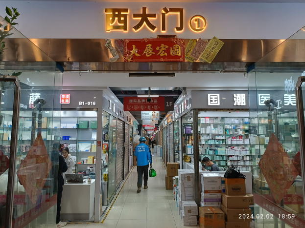 Ein Blick in eine chinesische Apotheke mit Glaswänden, Regalen voller Gegenstände, verstreuten Kartons und einer Pflanze links.