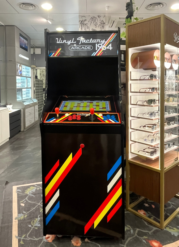 Ein Geschäft mit einem zentralen Retro-Arcade-Automaten, einem Gestell mit Gläsern auf der rechten Seite und Hintergrund-Elementen wie Schränken, einem Fernseher und anderen Gegenständen, mit Deckenleuchten, die eine retro-mäßige Atmosphäre schaffen.