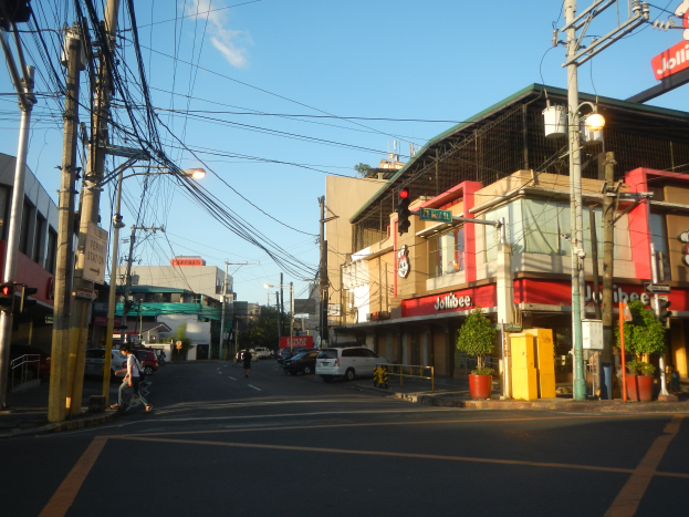 Eine Stadtstraße mit Autos, Fußgängern, Strommasten, Verkehrszeichen, Gebäuden mit Schildern, Topfpflanzen und einem bewölkten Himmel.