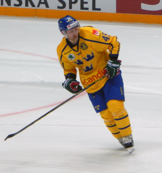 Eishockeyspieler in Bewegung auf dem Eis, mit Schutzausrüstung und Stock, mit einer Tafel im Hintergrund, auf der Text steht.