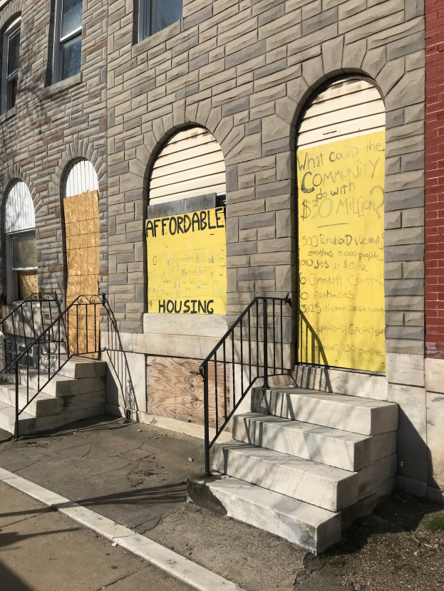 Ein zweistöckiges Gebäude mit vernagelten Fenstern und einem verblassten "Günstiger Wohnraum"-Schild, das über eine Treppe mit Geländern erreichbar ist und neben einer Straße steht.