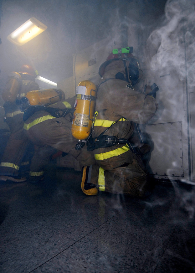 Feuerwehr in einem raucherfüllten Raum mit beleuchteter Ausrüstung und einer Wand im Hintergrund.