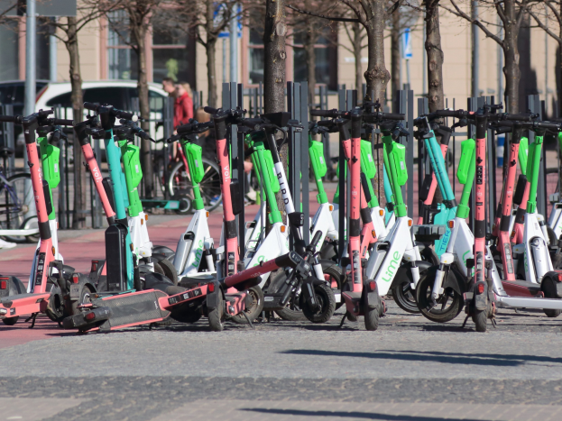 Eine Reihe von Elektro-Rollern, die an einer Straße geparkt sind, mit einer Person im Vordergrund, Bäumen, Masten, Schildern, Fahrzeugen und Gebäuden im Hintergrund.