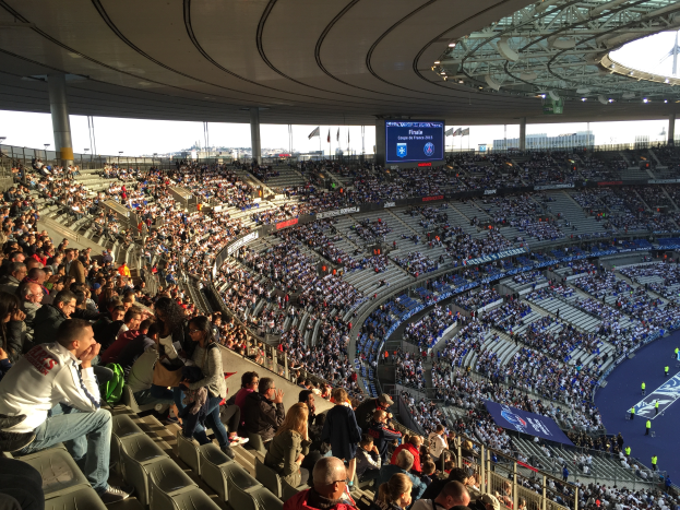Eine große Menschenmenge sitzt im Allianz Arena in München, Deutschland, bei einem Fürballspiel, mit einer Bühne auf der rechten Seite, Fahnen, Masten und einem Bildschirm im Hintergrund und der Himmel oben sichtbar.