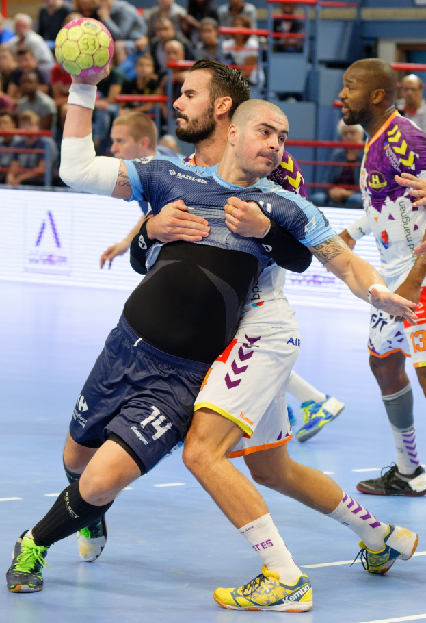 Eine Gruppe von Männern, die Handball spielen, mit einem Spieler, der den Ball hält, und Zuschauern im Hintergrund.