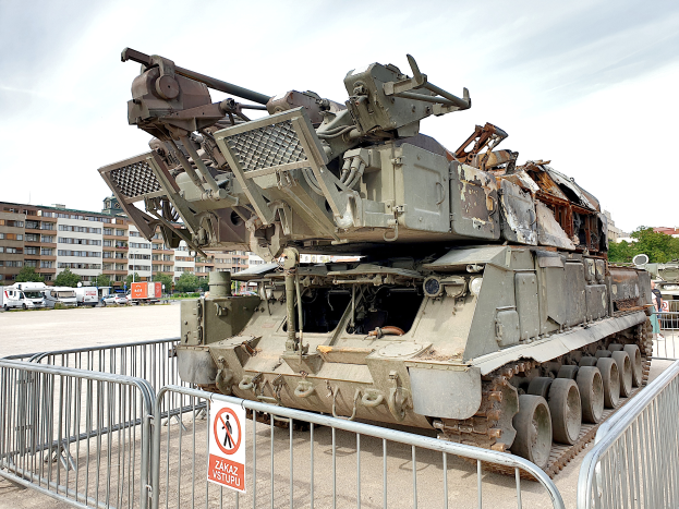 Ein gepanzertes Fahrzeug steht vor einem Metallzaun mit einem Schild, dahinter sind Fahrzeuge, Bäume, Gebäude und ein klarer blauer Himmel zu sehen.