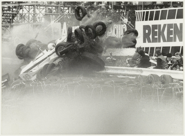 Ein Schwarz-Weiß-Foto eines verunglückten Rennwagens umgeben von Reifen, mit einer Gruppe von Menschen rechts und einer Tafel mit Text im Hintergrund.
