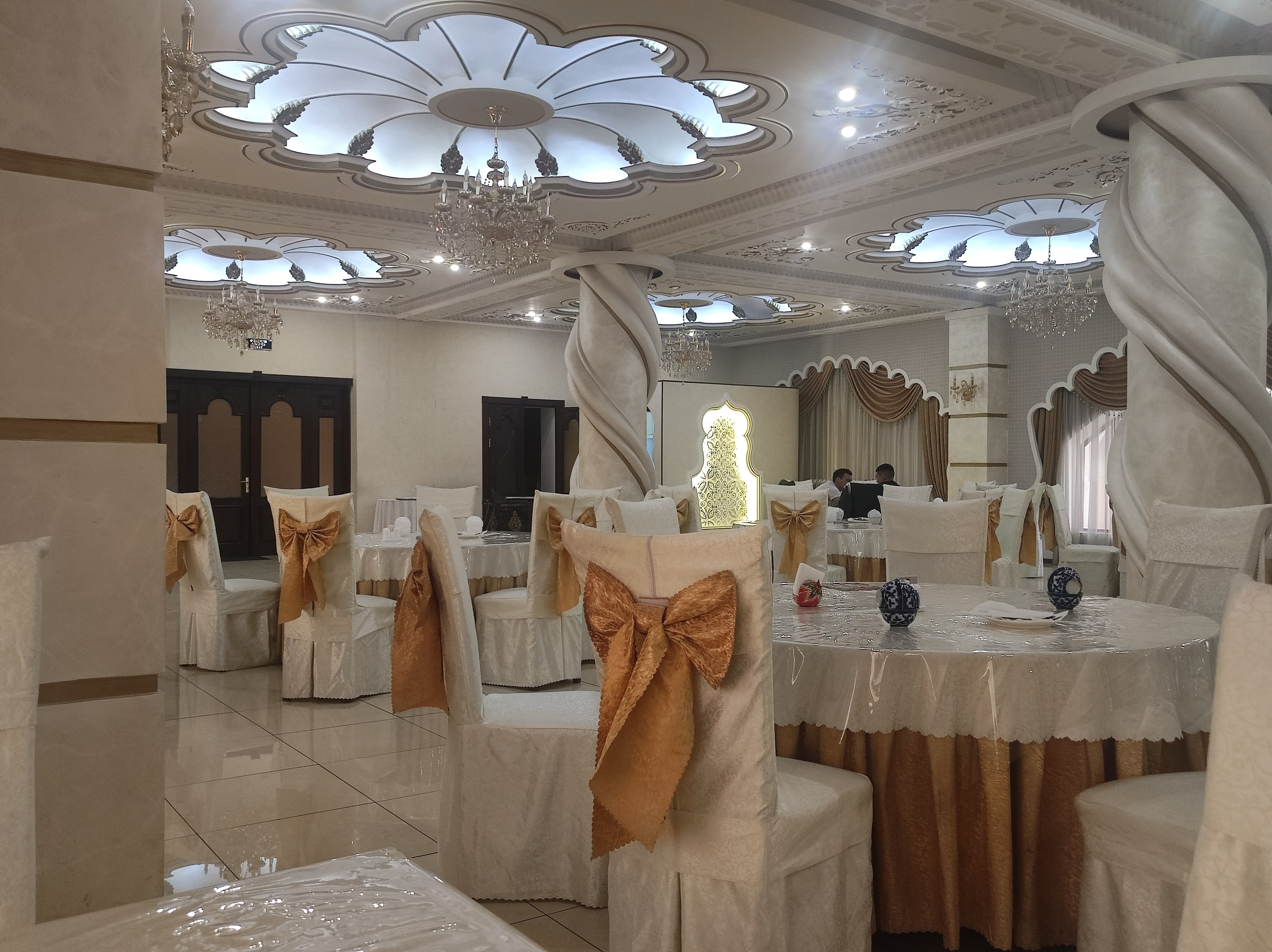 Ein großer Festsaal mit Tischen und Stühlen, die mit Tüchern drapiert und mit verschiedenen Dekorationen geschmückt sind, mit Wänden, Türen und Vorhängen sowie einer Decke mit Lichtern und Kronleuchtern.