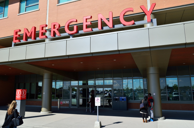 Eine Gruppe von Menschen vor einem Glaswand-Notfallgebäude mit Säulen, einem Namensschild, Hinweistafeln auf Pfählen und einem Müllcontainer.