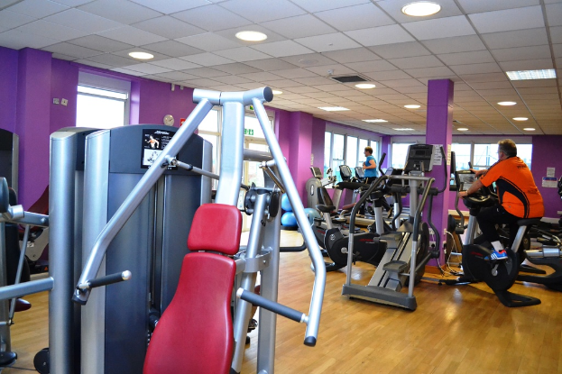 Ein Fitnessstudio mit mehreren Geräten und einigen Menschen, die trainieren, mit einer Decke und Lampen oben im Bild.