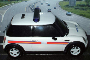 Ein weißer Polizeiwagen ist im Vordergrund geparkt, hinter ihm bewegen sich mehrere Fahrzeuge auf der Straße, umgeben von Gras und Bäumen auf beiden Seiten.