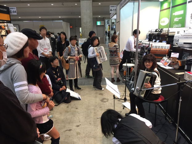 Eine Gruppe von Menschen spielt Musikinstrumente um einen Tisch, mit einer Frau, die in der Mitte auf einem Stuhl eine Akkordeon spielt; ein Tisch mit Gegenst├Ąnden ist rechts und eine S├Ąule, Texttafeln und Deckenleuchten sind im Hintergrund bei einem japanischen Musikfestival zu sehen.