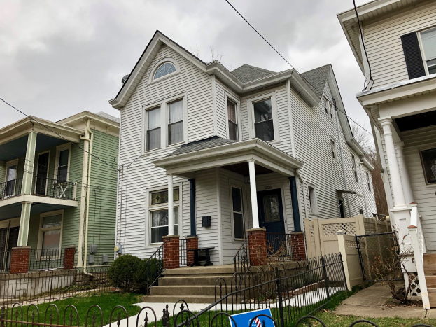 Reihe von Häusern mit Fenstern, Türen, Säulen, Geländern, Stufen und Dächern, umgeben von einem Metallzaun, Gras, Pflanzen, Bäumen und einem bewölkten Himmel, mit einer Mietanzeige am unteren Rand, die die Adresse und Listen Details anzeigt.